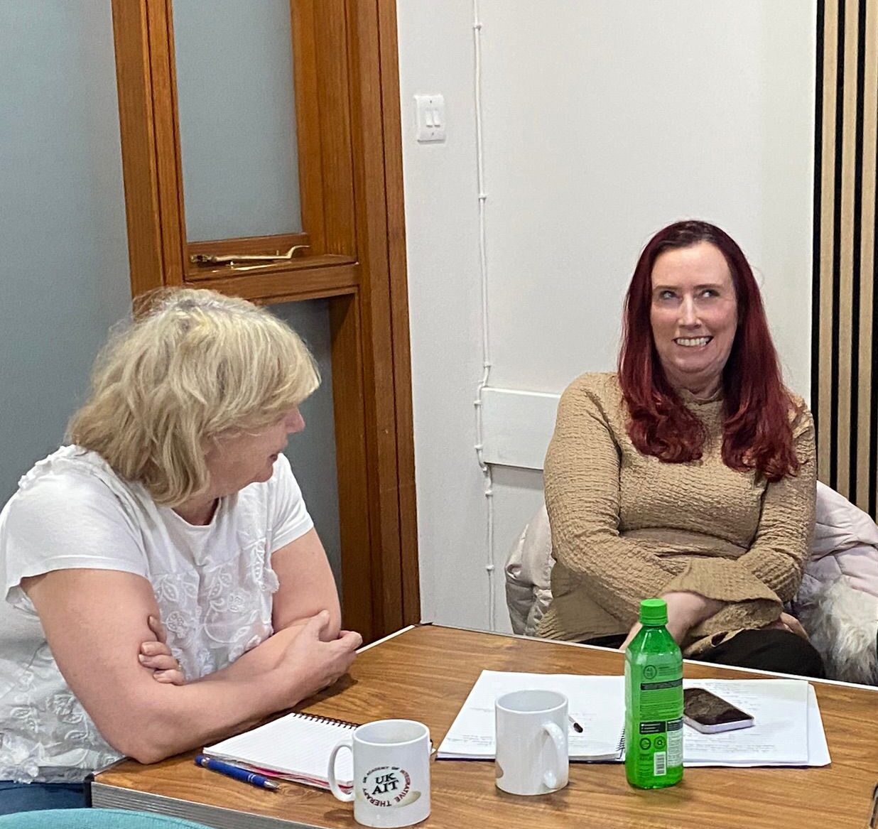 Two women having a conversation at a table with notebooks, mugs, and a green bottle.