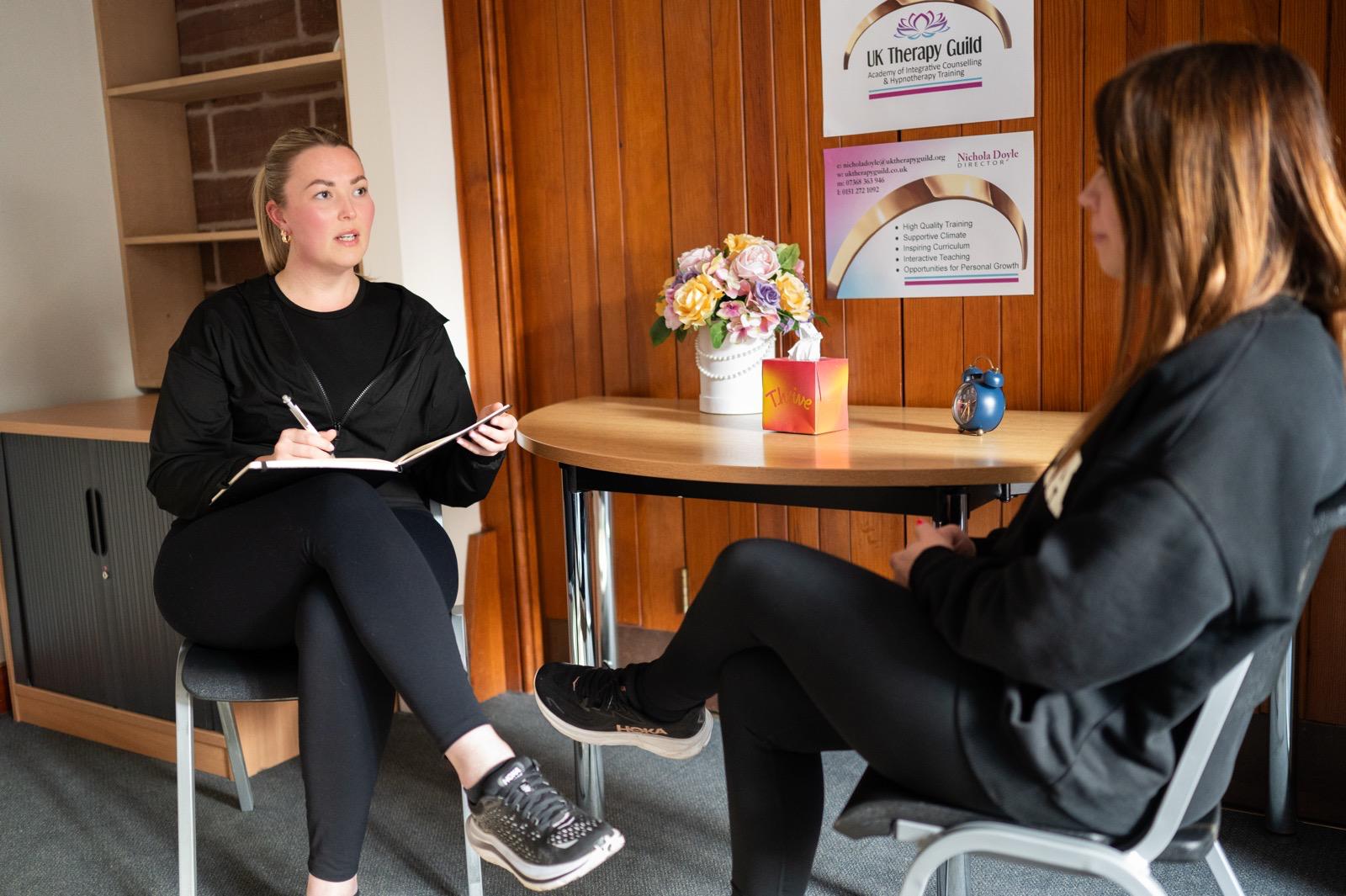 Counselling training session: tutor taking notes with client in Liverpool teaching room, flowers and UK Therapy Guild sign