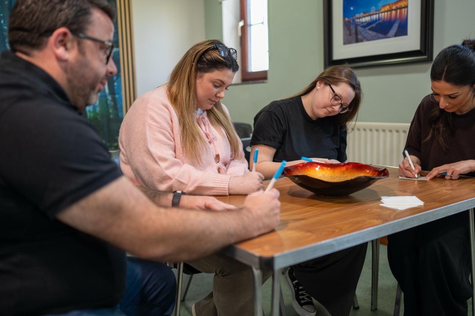 Counselling students in Liverpool workshop writing notes at a wooden table around a decorative bowl, classroom training setting.