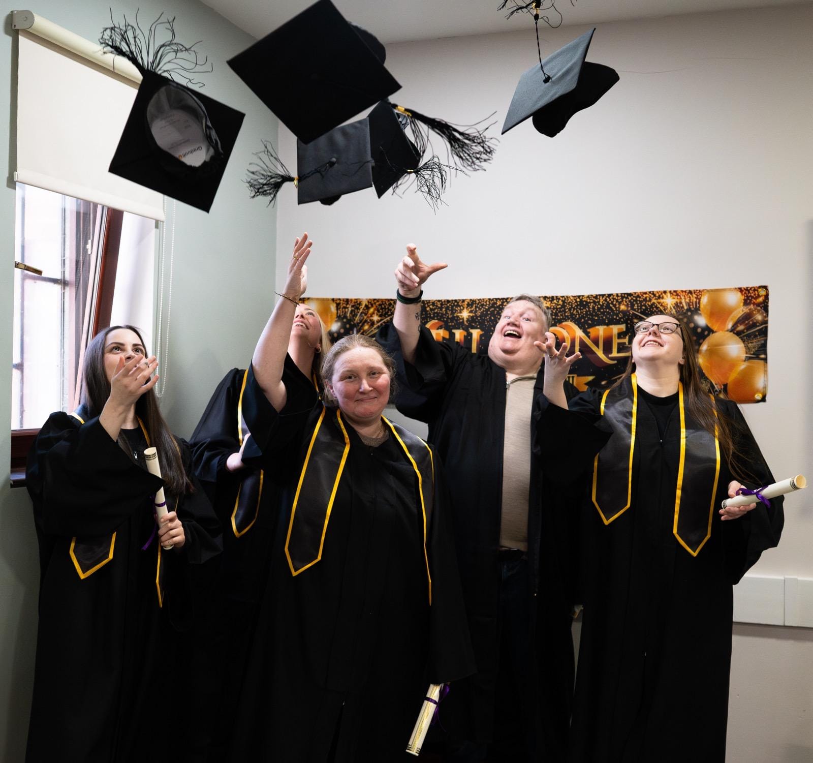 Trainee counsellors in black robes with gold trim toss mortarboards, holding diplomas at a Liverpool therapy graduation.