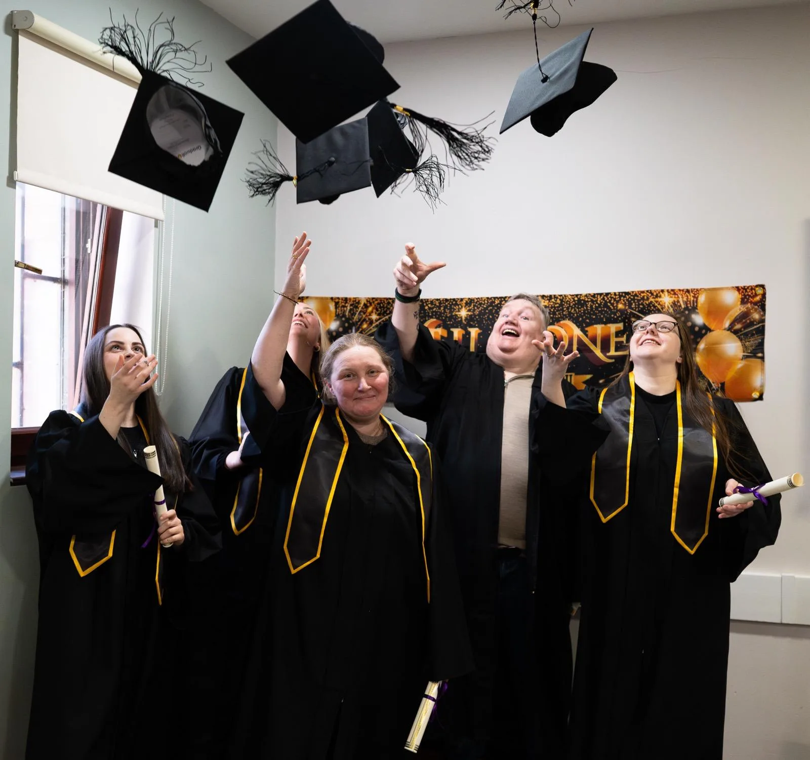 Trainee counsellors in black robes with gold trim toss mortarboards, holding diplomas at a Liverpool therapy graduation.