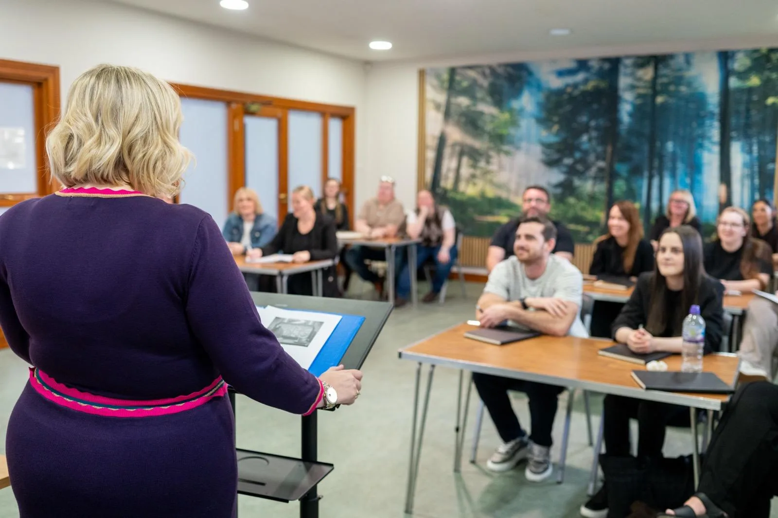 Tutor in purple at a lectern leading an adult counselling training session in a Liverpool classroom with a forest mural backdrop