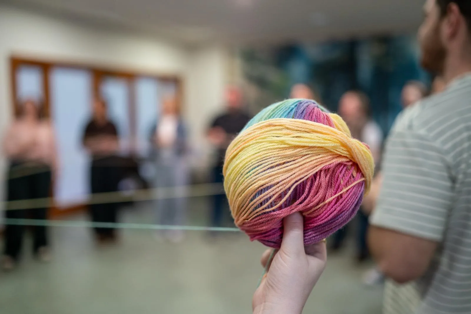 Hand holding rainbow yarn ball with string linking a blurred group in a circle — counselling training exercise, Liverpool.