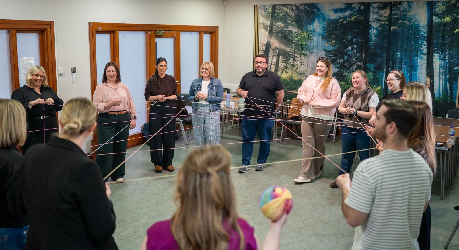 Therapy trainees standing in a circle creating a web with coloured yarn in a classroom with forest mural backdrop, Liverpool