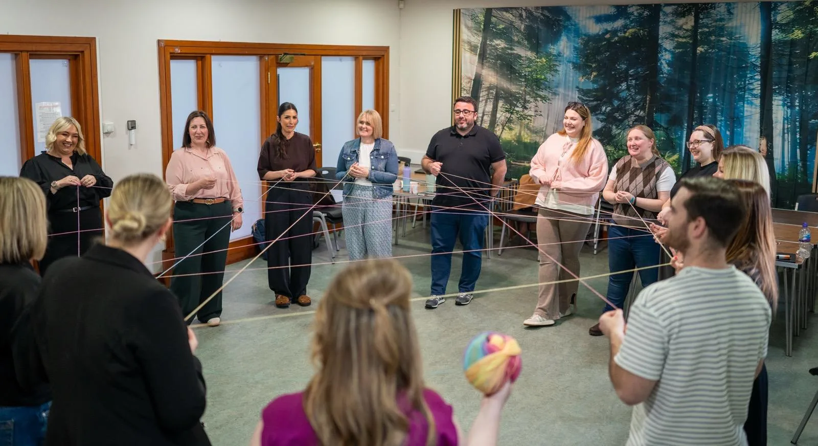 Therapy trainees standing in a circle creating a web with coloured yarn in a classroom with forest mural backdrop, Liverpool